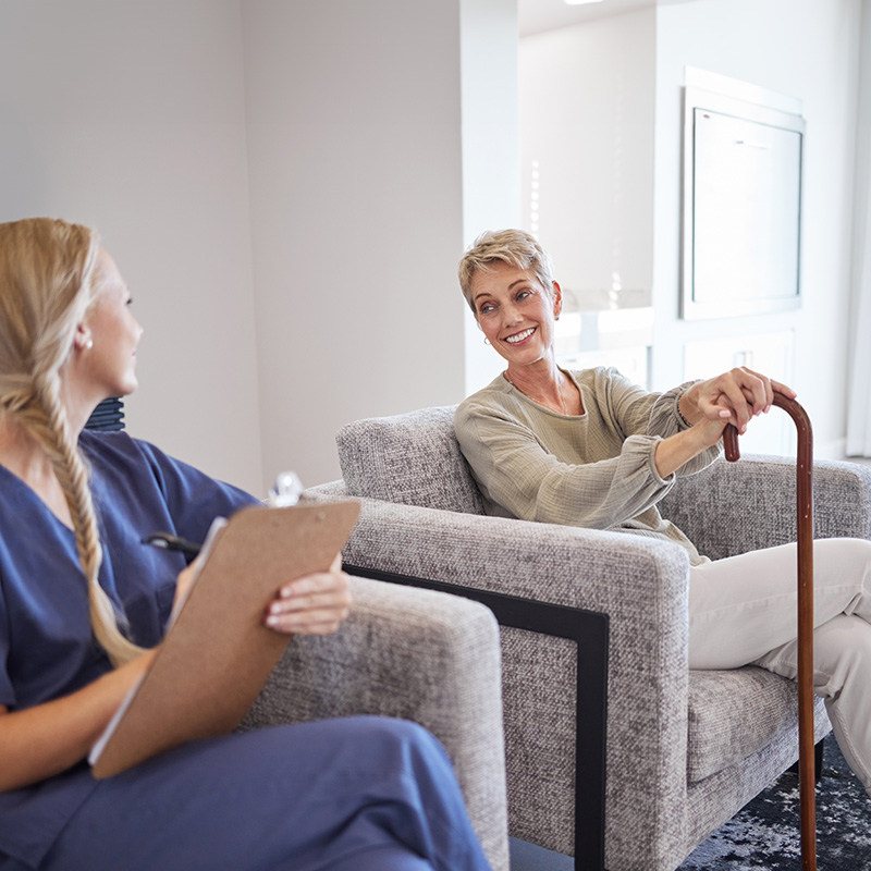 An elderly woman in a gray sweater is seated on a couch, engaging with a nurse holding a clipboard.