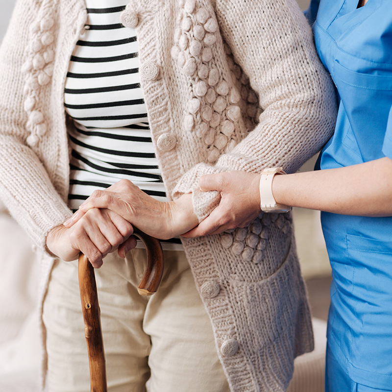 An elderly woman in a beige cardigan being assisted by a healthcare professional, likely a nurse or caregiver, with a walker during what appears to be a medical visit.